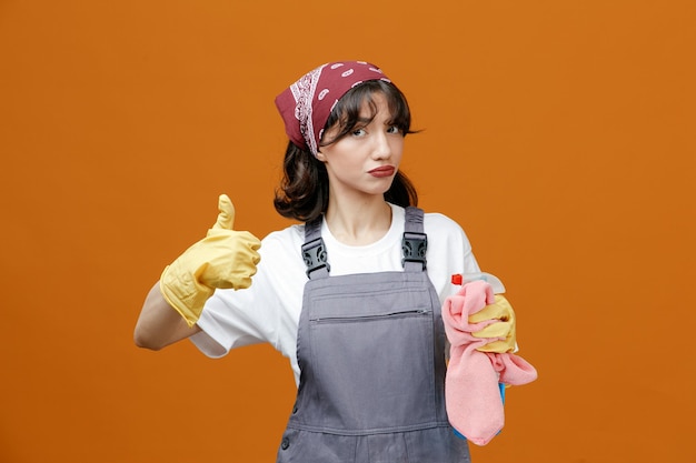 Confident young female cleaner wearing uniform rubber gloves and bandana holding cleanser and cloth duster looking at camera showing thumb up isolated on orange background