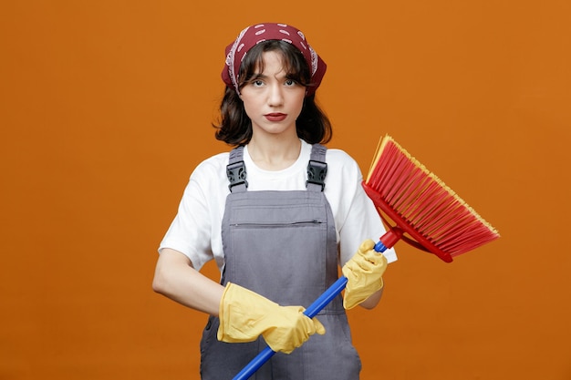 Уверенная молодая женщина-уборщица в резиновых перчатках и бандане, держащая швабру обеими руками и смотрящая в камеру на оранжевом фоне