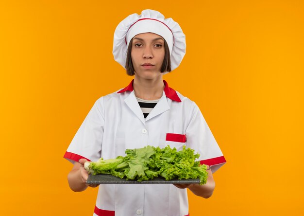 Confident young female cook in chef uniform holding cutting board with lettuce on it and looking isolated on orange  with copy space