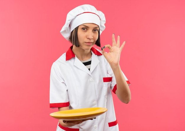 Confident young female cook in chef uniform holding empty plate and doing ok sign isolated on pink  with copy space