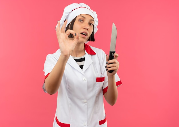 Free photo confident young female cook in chef uniform holding knife and doing ok sign isolated on pink  with copy space