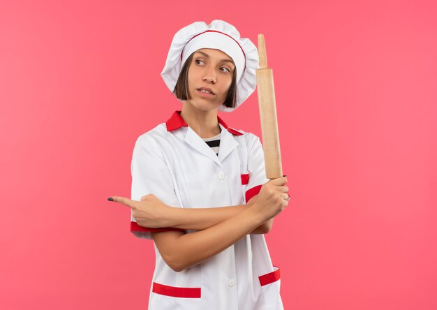 Confident young female cook in chef uniform holding rolling pin and looking and pointing at side isolated on pink  with copy space