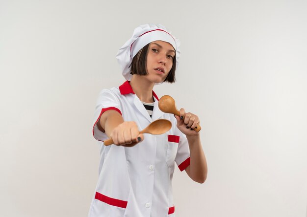 Confident young female cook in chef uniform holding and stretching out spoons isolated on white  with copy space