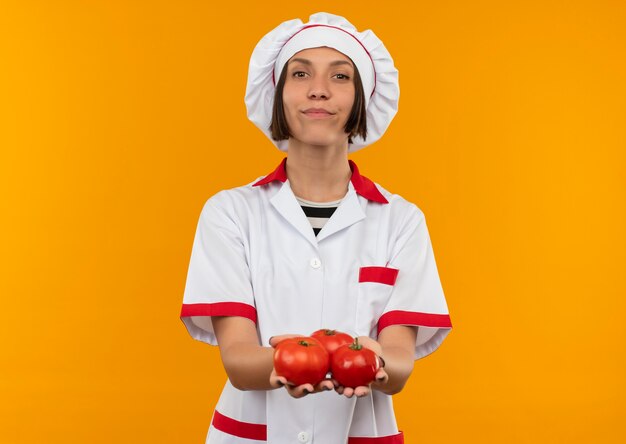 Уверенная молодая женщина-повар в униформе шеф-повара держит помидоры, изолированные на апельсине, с копией пространства