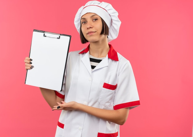 Confident young female cook in chef uniform showing and pointing with hand at clipboard isolated on pink  with copy space