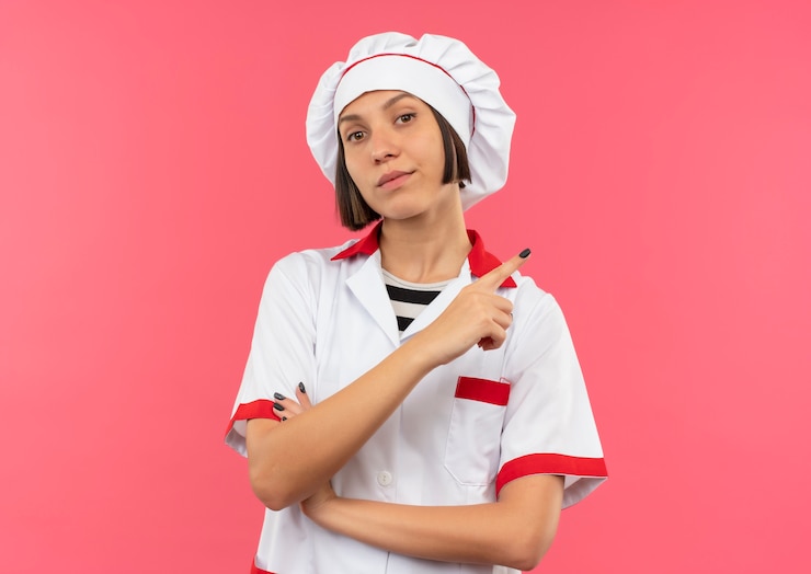 Free Photo confident young female cook in chef uniform standing with closed posture and pointing at side isolated on pink  with copy space