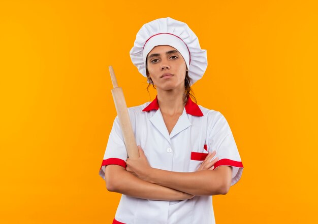 Confident young female cook wearing chef uniform crossing hands and holding rolling pin on isolated yellow wall with copy space
