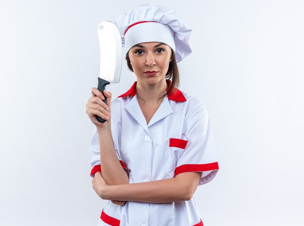 confident young female cook wearing chef uniform holding cleaver isolated on white wall