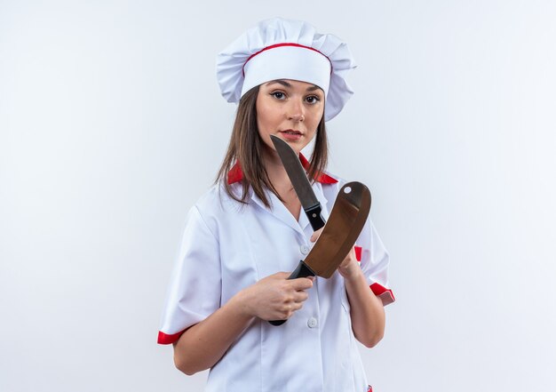 confident young female cook wearing chef uniform holding and crossing cleaver with knife isolated on white wall