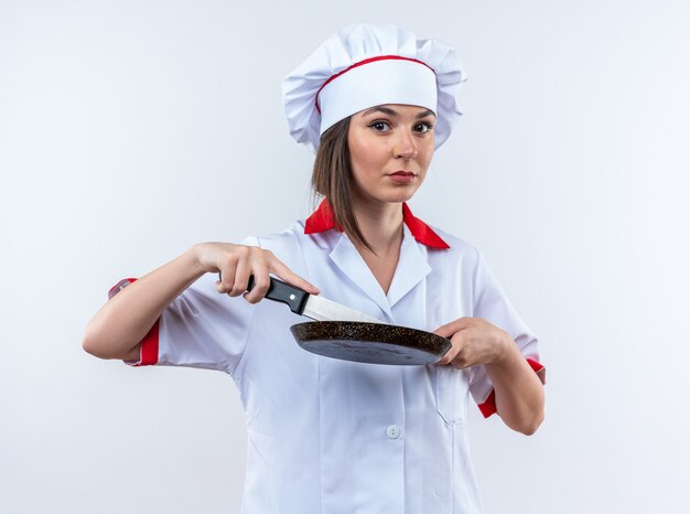 흰색 배경에 고립 된 칼으로 프라이팬을 들고 요리사 유니폼을 입고 자신감 젊은 여성 요리사