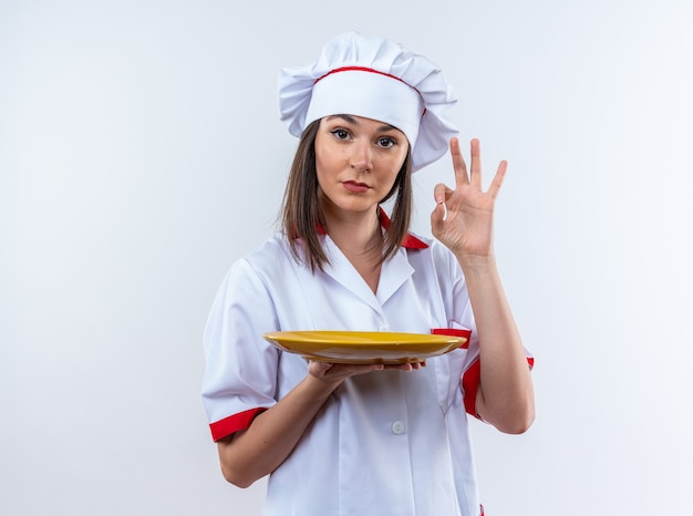 Confident young female cook wearing chef uniform holding plate showing okay gesture isolated on white wall