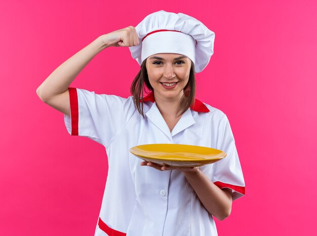 confident young female cook wearing chef uniform holding plate showing strong gesture isolated on pink wall