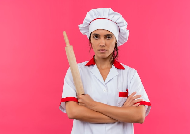 Уверенная молодая женщина-повар в униформе шеф-повара держит скалку и скрещивает руки на изолированной розовой стене с копией пространства