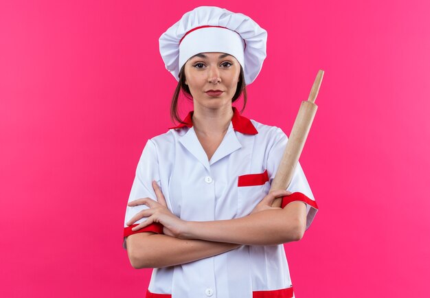 confident young female cook wearing chef uniform holding rolling pin crossing hands isolated on pink wall