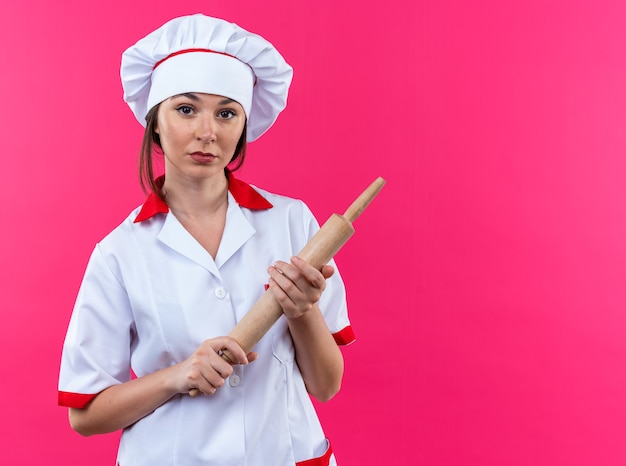 복사 공간 분홍색 배경에 고립 된 롤링 핀을 들고 요리사 유니폼을 입고 자신감 젊은 여성 요리사
