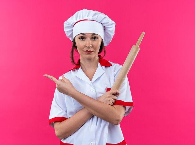 Confident young female cook wearing chef uniform holding rolling pin points at side isolated on pink background with copy space