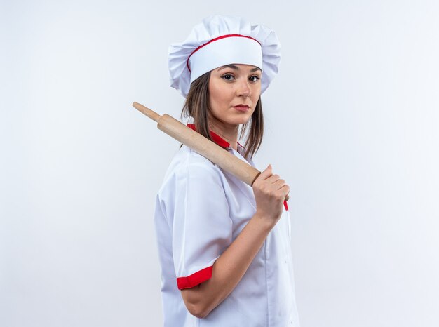 Уверенная молодая женщина-повар в униформе шеф-повара держит скалку на плече, изолированную на белой стене