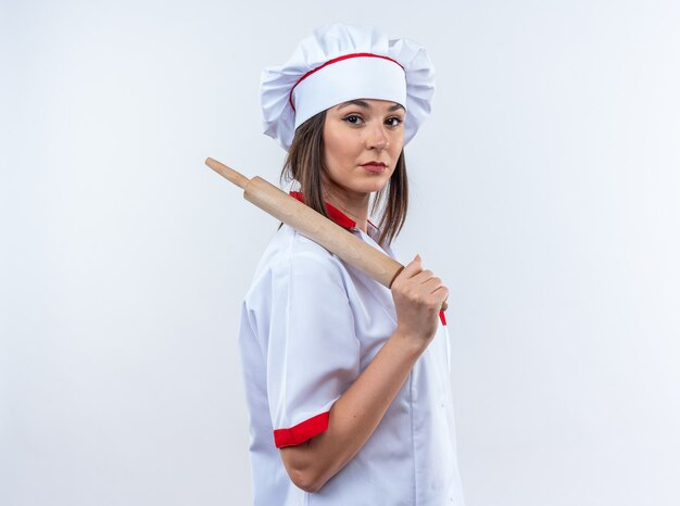 Confident young female cook wearing chef uniform holding rolling pin on shoulder isolated on white wall