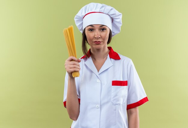 confident young female cook wearing chef uniform holding spaghetti isolated on olive green wall