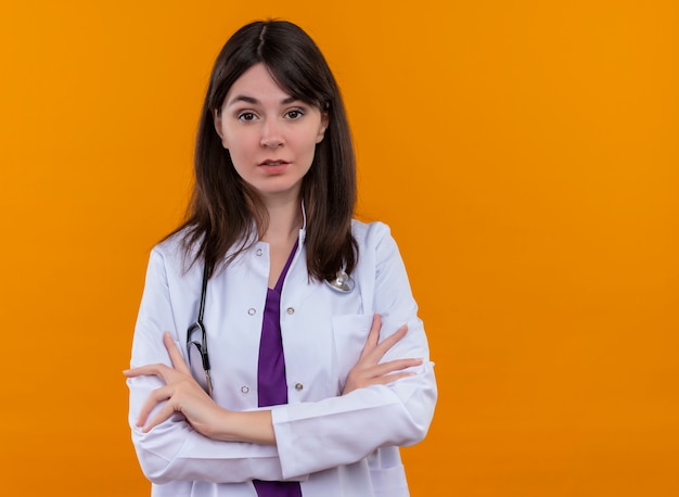 Confident young female doctor in medical robe with stethoscope crosses arms on isolated orange background with copy space