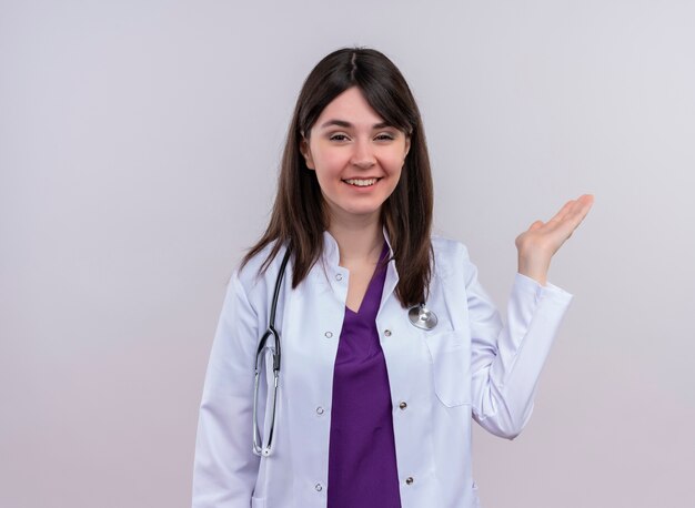 Confident young female doctor in medical robe with stethoscope holds empty hand up on isolated white background with copy space