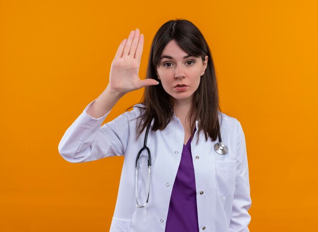 Confident young female doctor in medical robe with stethoscope holds hand up on isolated orange background with copy space