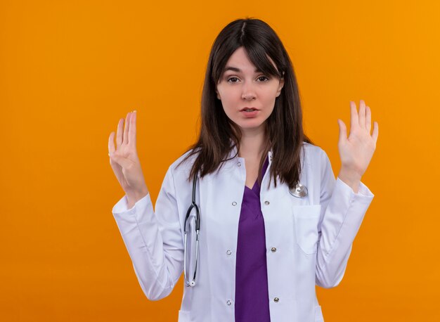 Confident young female doctor in medical robe with stethoscope holds hands open on isolated orange background with copy space