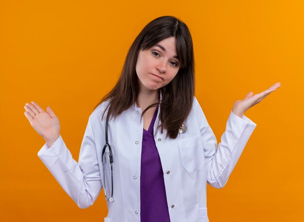 Confident young female doctor in medical robe with stethoscope holds hands up on isolated orange background