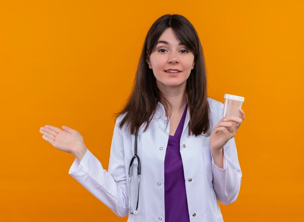 Confident young female doctor in medical robe with stethoscope holds medicine cup on isolated orange background with copy space