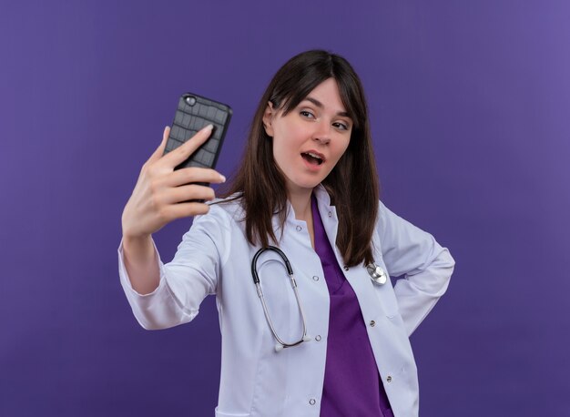 Confident young female doctor in medical robe with stethoscope holds phone and looks at phone on isolated violet background with copy space