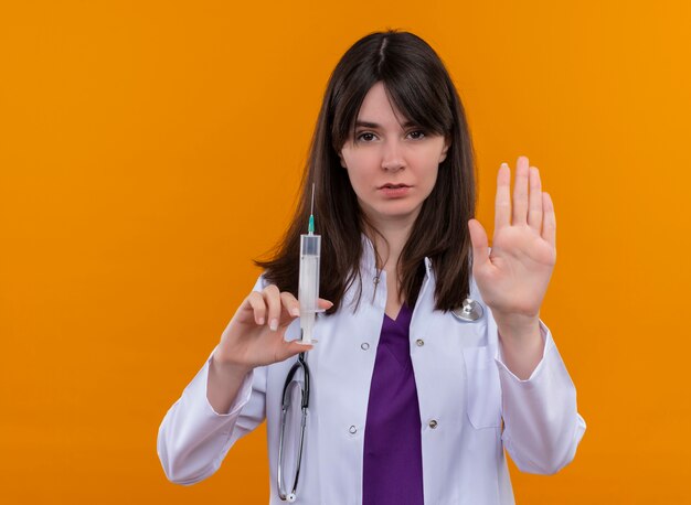 Уверенная молодая женщина-врач в медицинском халате со стетоскопом держит шприц и останавливает жесты на изолированном оранжевом фоне