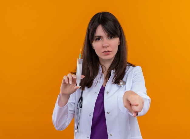 Уверенная молодая женщина-врач в медицинском халате со стетоскопом держит шприц и указывает на камеру на изолированном оранжевом фоне с копией пространства
