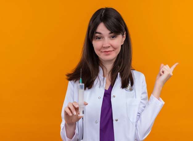 Confident young female doctor in medical robe with stethoscope holds syringe and points to the side on isolated orange background with copy space 0