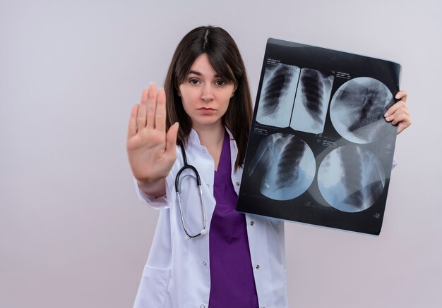 Confident young female doctor in medical robe with stethoscope holds x-ray shot and gestures stop on isolated white background with copy space