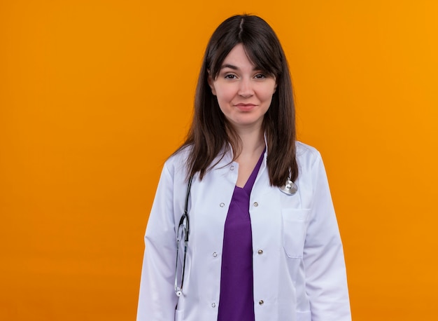 Confident young female doctor in medical robe with stethoscope looks at camera on isolated orange background with copy space