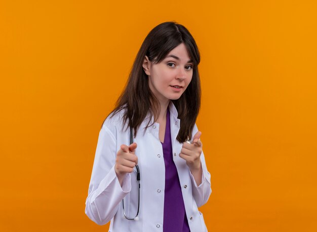 Confident young female doctor in medical robe with stethoscope points at camera with both hands on isolated orange background with copy space