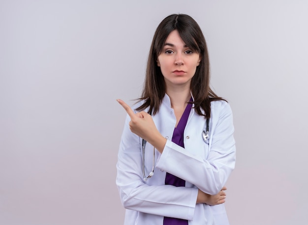 Confident young female doctor in medical robe with stethoscope points to the side and looks at camera on isolated white background with copy space