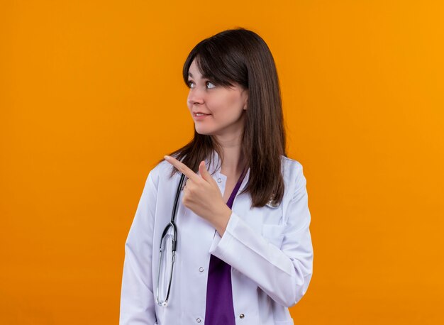 Confident young female doctor in medical robe with stethoscope points to the side and looks to the side on isolated orange background with copy space