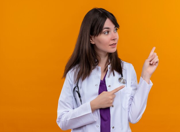 Confident young female doctor in medical robe with stethoscope points to the side with both hands on isolated orange background with copy space