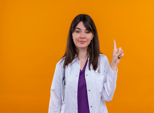 Confident young female doctor in medical robe with stethoscope points up on isolated orange background with copy space