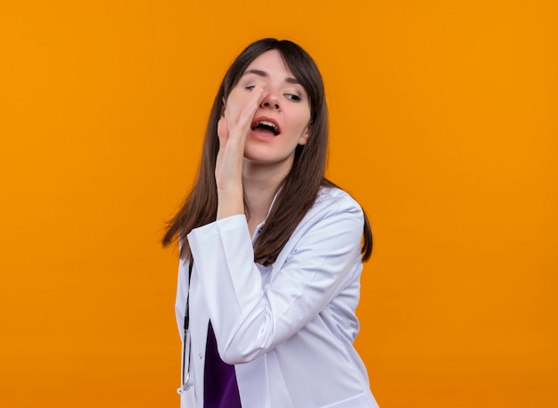 Confident young female doctor in medical robe with stethoscope pretends to call someone on isolated orange background with copy space