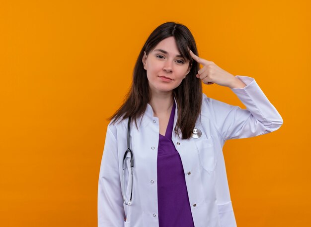Confident young female doctor in medical robe with stethoscope puts finger on her head on isolated orange background with copy space