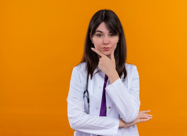 Confident young female doctor in medical robe with stethoscope puts hand on chin on isolated orange background with copy space