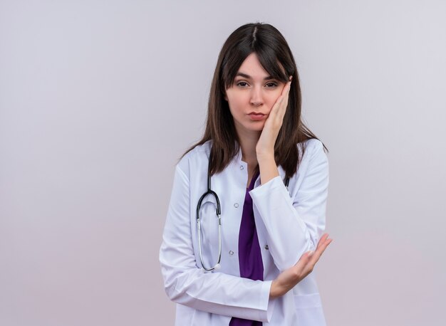 Confident young female doctor in medical robe with stethoscope puts hand on chin on isolated white background with copy space