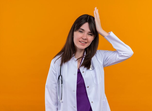 Confident young female doctor in medical robe with stethoscope puts hand on head on isolated orange background with copy space