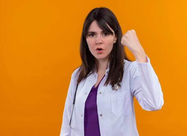 Confident young female doctor in medical robe with stethoscope raises fist on isolated orange background with copy space