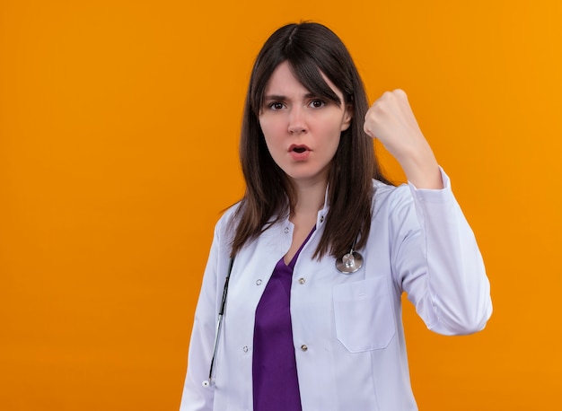 Уверенная молодая женщина-врач в медицинском халате со стетоскопом поднимает кулак на изолированном оранжевом фоне с копией пространства