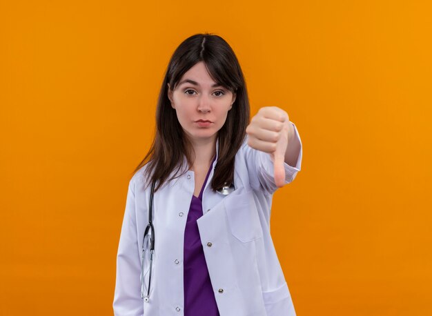 Confident young female doctor in medical robe with stethoscope thumbs down on isolated orange background with copy space