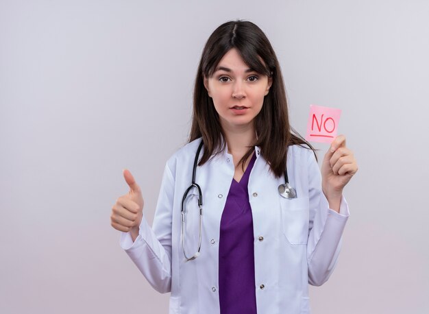 Confident young female doctor in medical robe with stethoscope thumbs up and holds no on isolated white background with copy space