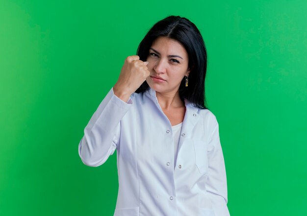 Confident young female doctor wearing medical robe  doing be strong gesture isolated on green wall with copy space